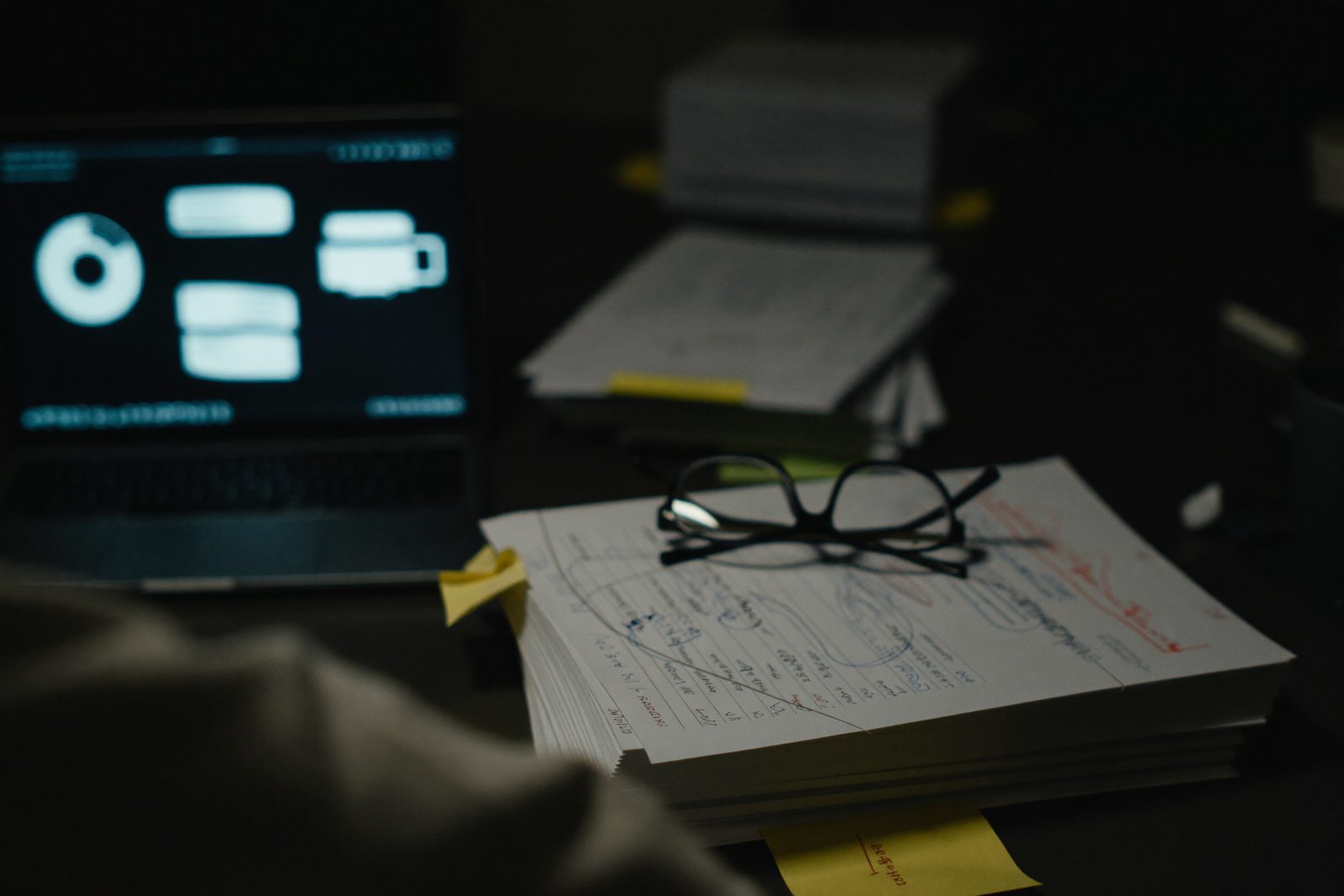 A desk at night: stacks of invoices and records beside a glowing laptop.